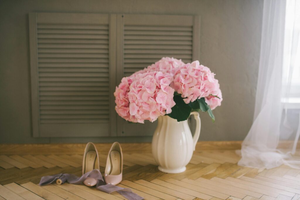 Pink hydrangeas in a vase with elegant bridal shoes on a wooden floor.