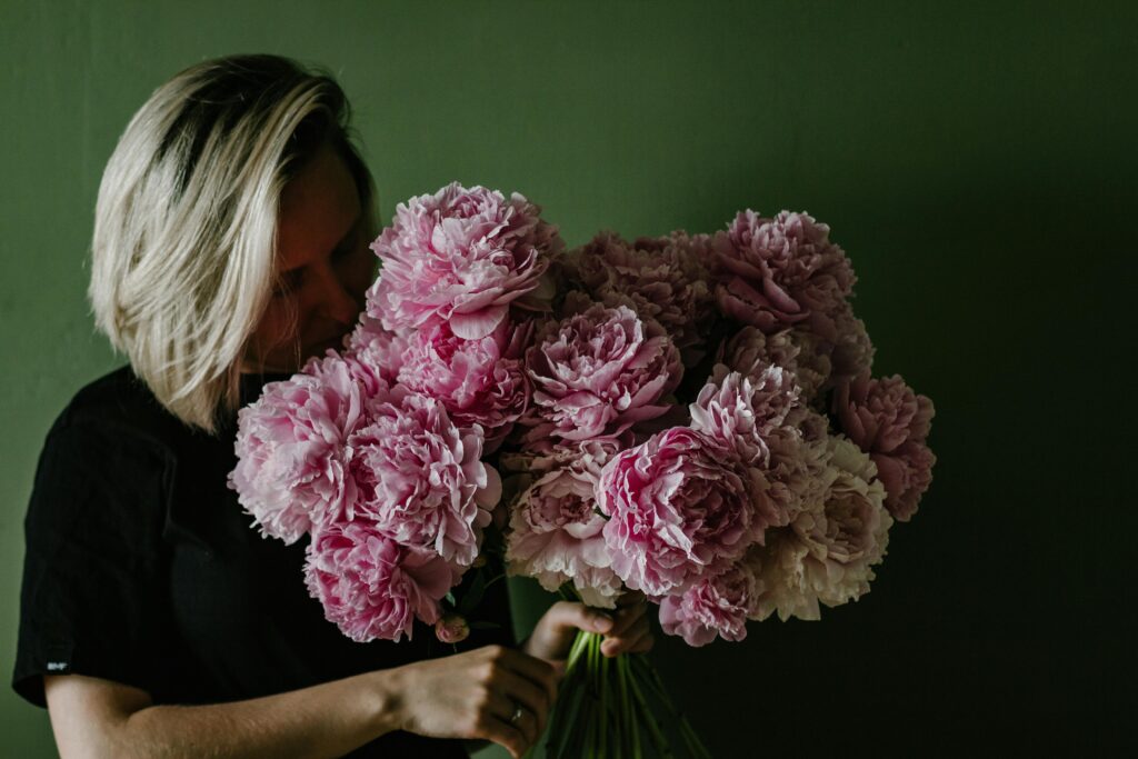 Female with dyed hair smelling bouquet of fragrant pink blooming peonies against green background