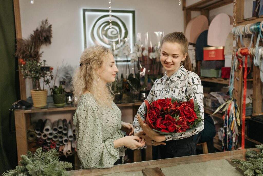 Two women happily exchange a vibrant red bouquet in a cozy flower shop setting.