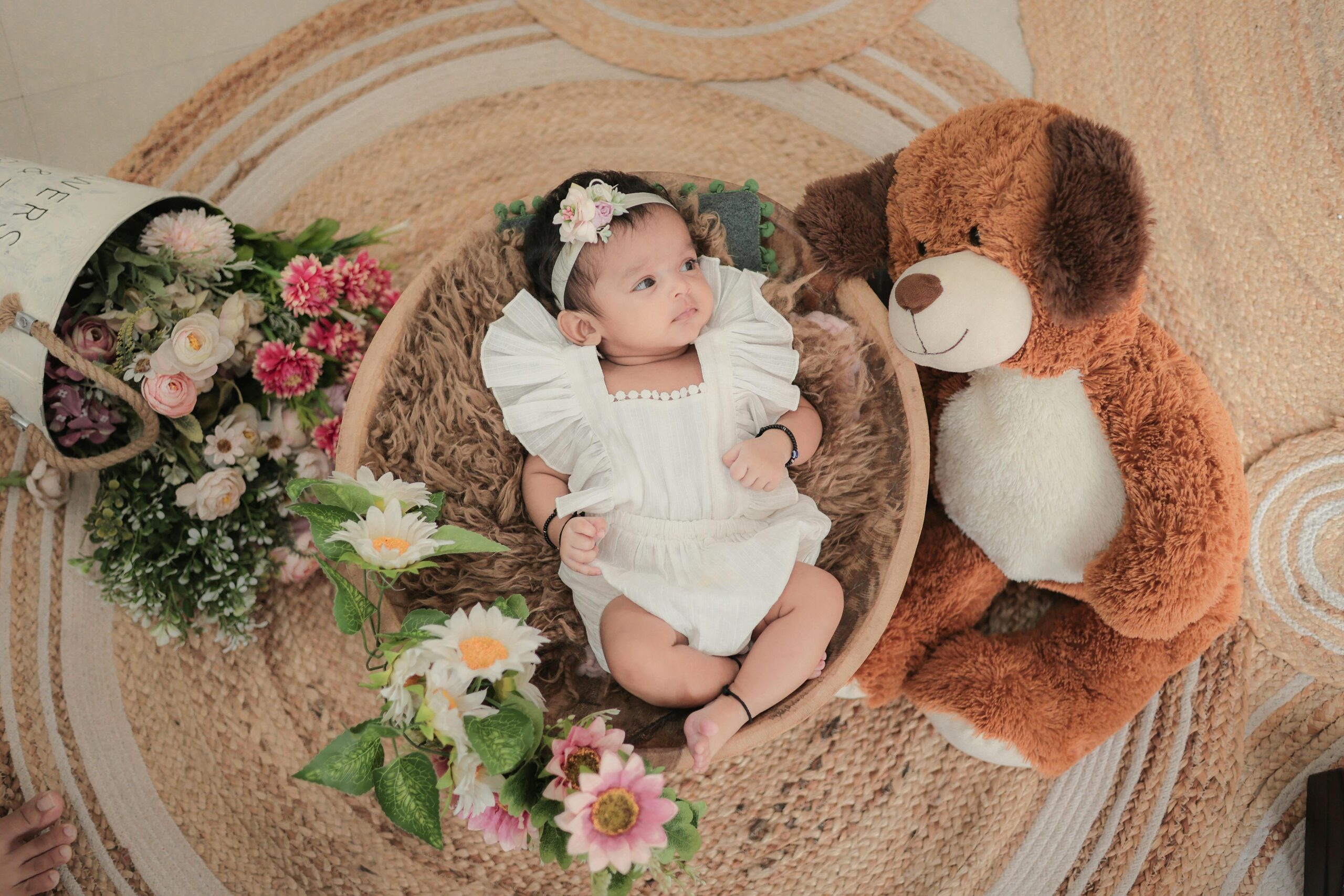 Charming scene of a baby lying in a basket surrounded by flowers and a teddy bear indoors.
