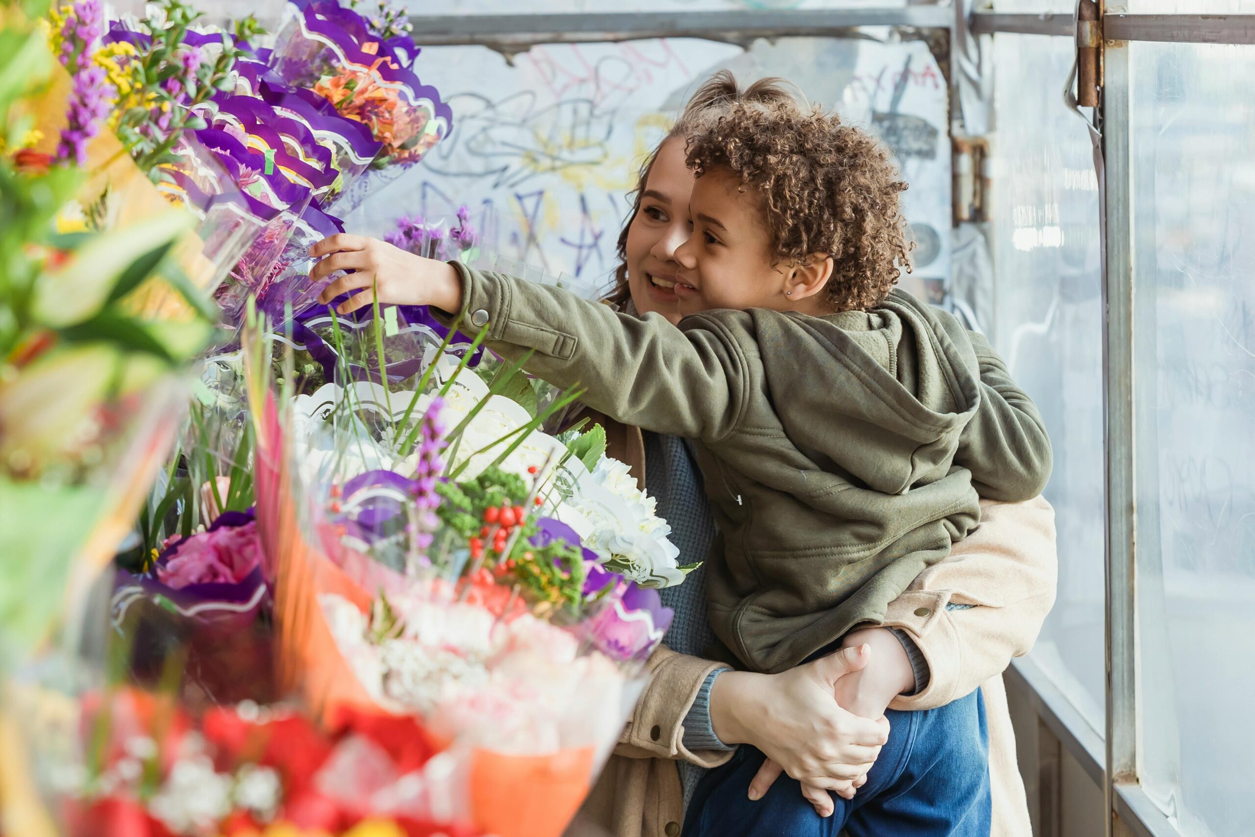 Smiling mother holding cute black son on hands while choosing together bouquet in floristry store